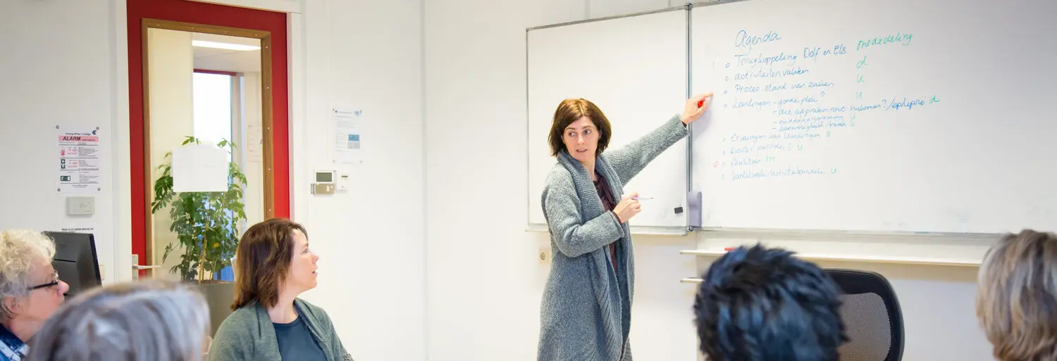 Een vrouw geeft voor een whiteboard een presentatie voor een groep mensen en bespreekt het Jaarverslag. School en Veiligheid