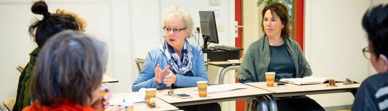 Een groep mensen die in een klaslokaal aan een bureau zitten en begeleiding krijgen van een vertrouwenspersoon. School en Veiligheid