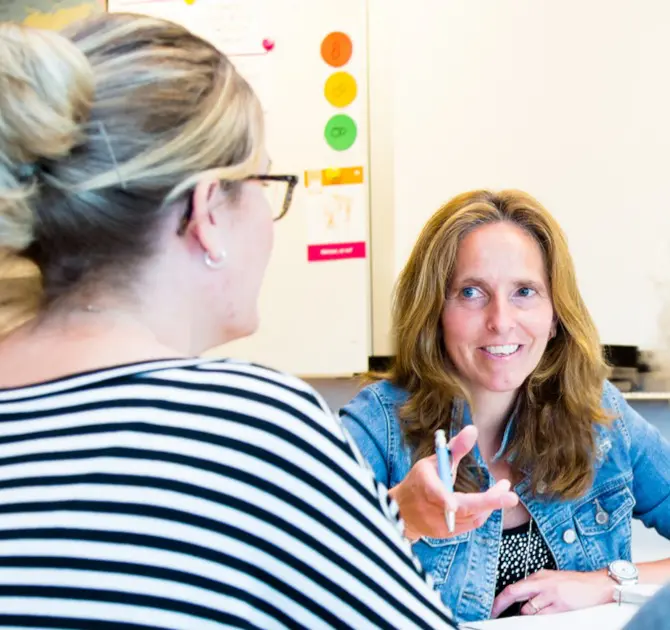 Twee vrouwen voerden een gesprek over het ondersteunen van scholen met sociale veiligheid, waarbij één een pen vasthield en gebaren maakte tijdens het spreken. School en Veiligheid