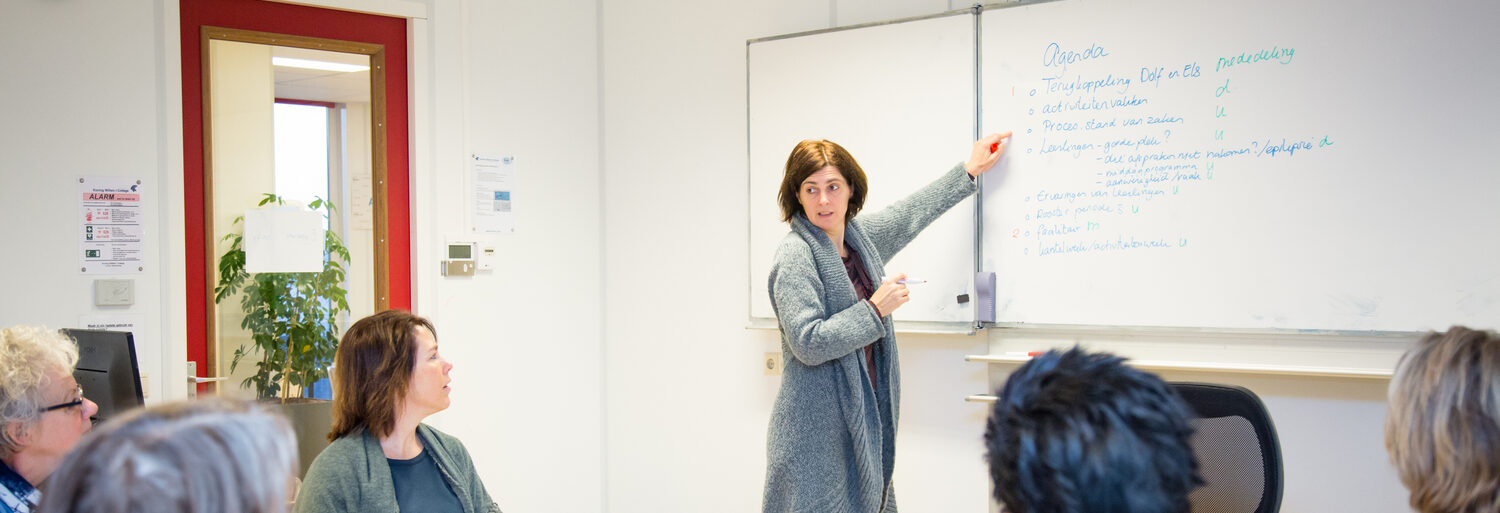 Een vrouw geeft voor een whiteboard een presentatie voor een groep mensen en bespreekt het Jaarverslag. School en Veiligheid