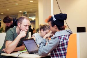 Een groep mensen die met laptops rond een tafel zitten en discussiëren over discriminatie in het voortgezette onderwijs. School en Veiligheid