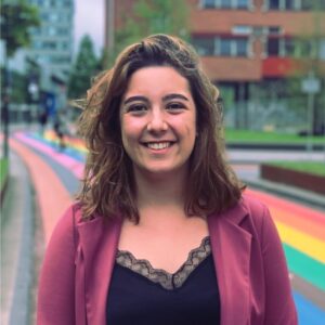 Loïs Gampierakis, een vrouw, glimlachend voor een regenboogkleurige straat. School en Veiligheid