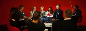Een diverse groep mensen zittend op stoelen voor een rode muur, die gender en seksuele diversiteit in het mbo vertegenwoordigen. School en Veiligheid