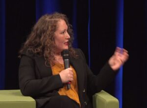 Een vrouw die deelneemt aan een onderwijsconferentie, zittend op een stoel en met alle respect in een microfoon spreekt. School en Veiligheid