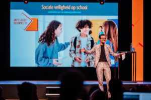 Een presentator in patroonpak gebaart energiek op de Onderwijsconferentie Met Alle Respect! over sociale veiligheid op scholen, met op de achtergrond een slideshow van lachende leerlingen School en Veiligheid