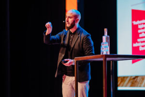 Een kale man in een donker jasje die presenteert met een afstandsbediening in de hand, staande naast een scherm met tekst op de "Onderwijsconferentie Met Alle Respect!" conferentie. School en Veiligheid