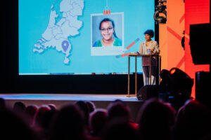 Een vrouw presenteert op de "Onderwijsconferentie Met Alle Respect!" met een digitaal scherm met een kaart en een portret achter haar, voor een aandachtig publiek. School en Veiligheid