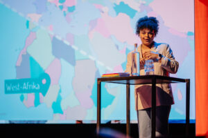 Op een podium van de Onderwijsconferentie Met Alle Respect! staat een persoon met blauw haar, sprekend en gebarend, met een kaart van West-Afrika en een waterfles ernaast School en Veiligheid