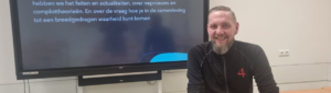 Glimlachende man zittend aan een tafel voor een televisie met tekst over spanning en discussie in de klas. School en Veiligheid
