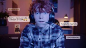 Een persoon met krullend haar, een geruit overhemd en een koptelefoon, kijkt in de camera. Meerdere chatbubbels met berichten in het Nederlands, inclusief opmerkingen en emoji's, zweven om hen heen. Een van hen noemt Week Tegen Pesten, wat het belang van vriendelijkheid benadrukt. School en Veiligheid