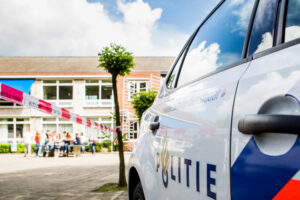Een politieauto staat geparkeerd vlakbij een plaats delict, afgeplakt met rood-wit politietape. Mensen verzamelen zich in de verte bij een gebouw van twee verdiepingen dat scholen ondersteunt bij initiatieven op het gebied van sociale veiligheid. School en Veiligheid
