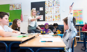 En juf samen met een groepje leerlingen van de basisschool. Een leerling steekt de hand op.