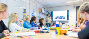 Groep mensen zit aan een tafel, een iemand staat voor de groep. Lijkt een overleg-setting.